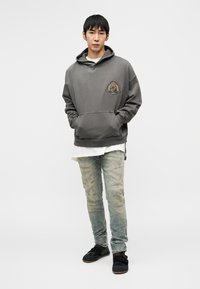 Jeune homme debout portant un sweat à capuche gris avec un emblème, un sous-maillot blanc, un jean délavé clair et des chaussures noires sur un fond blanc uni.