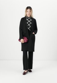 Nainen mustassa pitkässä bleiserissä ja housuissa, kuvioitua toppia pitäen, burgundinpunaisella clutch-laukulla, seisoo valkoista taustaa vasten.