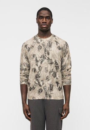 Mand iført en beige langærmet skjorte med sorte bladmønstre og grå bukser med sidelomme, stående foran en ensfarvet baggrund.