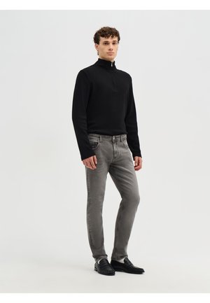 Giovane uomo in piedi che indossa un maglione nero a maniche lunghe con mezza cerniera, jeans grigi slim fit e scarpe slip-on nere su uno sfondo semplice.
