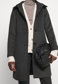 Szary wełniany płaszcz o strukturalnym fasonie, z kapturem i czarnymi guzikami. Nałożony na beżowy kardigan i szary golf; trzyma czarną torbę.