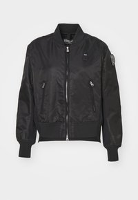 Černá bomber bunda s hladkým, lesklým materiálem, vroubkovanými manžetami a spodním lemem, předním zipem a dvěma bočními kapsami. Na rukávu se nachází nášivka.