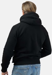 Czarna bluza z kapturem o luźnym kroju, z solidnym kapturem, ściągaczami i gładką fakturą. Noszona z jasnoniebieskimi dżinsami.