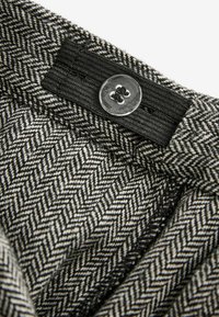 Grå och vit herringbone-tyg med en strukturerad yta, med ett svart elastiskt band och en rund metallknapp högst upp.