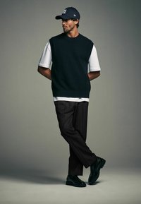 Dark green knitted sleeveless sweater over a white t-shirt, paired with black pinstriped trousers and green shoes, worn with a black cap.