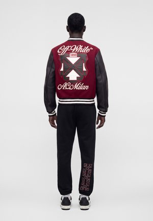 Muž s krátkymi vlasmi nosí červeno-čiernu varsity bundu Off-White a čierne nohavice s rovnajúcim sa logom, stojí chrbtom ku jednoliatemu pozadiu.