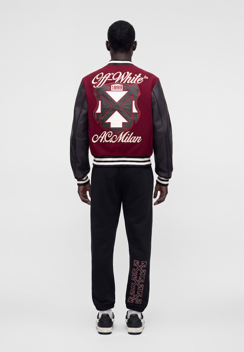 Homme aux cheveux courts portant une veste varsity Off-White rouge et noire et un pantalon noir avec un logo assorti, debout de dos devant un fond uni.
