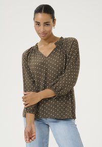 Blouse marron à pois avec un décolleté en V, encolure froncée et manches longues. Le tissu a un motif texturé, complété par une coupe décontractée.