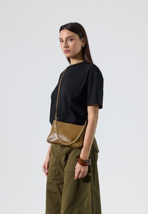 Joven mujer con camisa negra y pantalones verde oliva, lleva un bolso marrón cruzado con cadena dorada, usa pulseras marrones en la muñeca derecha.