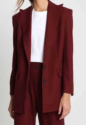 Femme portant un blazer bordeaux ajusté avec un pantalon taille haute assorti et un t-shirt blanc à col rond, posant devant un fond uni.