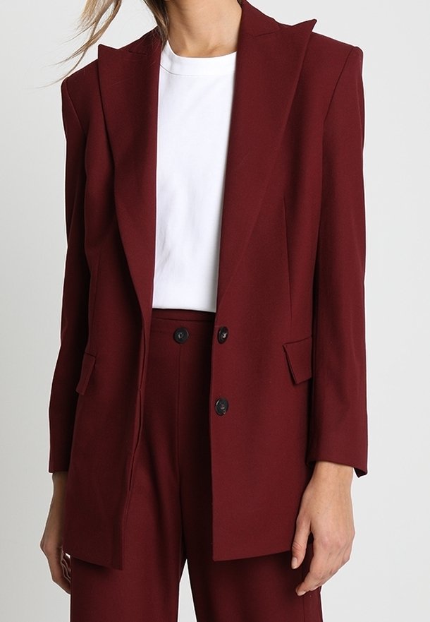 Femme portant un blazer bordeaux ajusté avec un pantalon taille haute assorti et un t-shirt blanc à col rond, posant devant un fond uni.