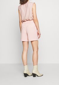 Top senza maniche di un rosa chiaro con rifiniture in pizzo, abbinato a pantaloni corti coordinati con una vita elastica. Il modello indossa stivaletti alla caviglia beige testurizzati.