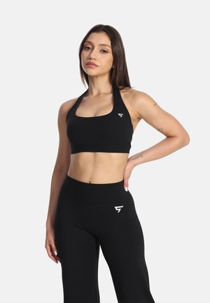 Junge Frau mit langen dunklen Haaren, die einen schwarzen Sport-BH und hoch taillierte schwarze Trainingsleggings mit Logos auf der linken Brust und am Oberschenkel trägt.