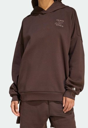 Brązowa, oversize bluza Adidas z napisem „three stripes studio” na piersi, noszona przez osobę wraz z dopasowanymi brązowymi szortami.