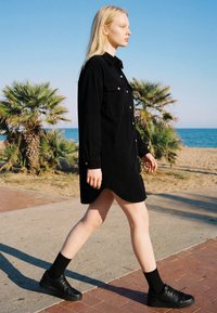 Blonde Frau trägt ein schwarzes Hemdkleid und Sneakers und läuft auf einem Strandweg mit Palmen und Ozean im Hintergrund.