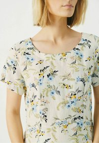Blusa de manga corta en tejido ligero que presenta un patrón floral con flores azules, amarillas y rosas sobre un fondo crema, con escote redondo.