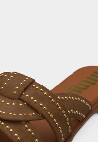 Sandale marron à lanières croisées ornées de petits clous dorés sur une semelle plate.