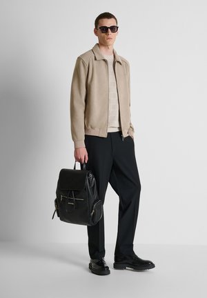 Mann trägt Sonnenbrille, beige Jacke, hellen Strickpullover, schwarze Hose, schwarze Schuhe, hält einen schwarzen Leder-Rucksack am Griff.