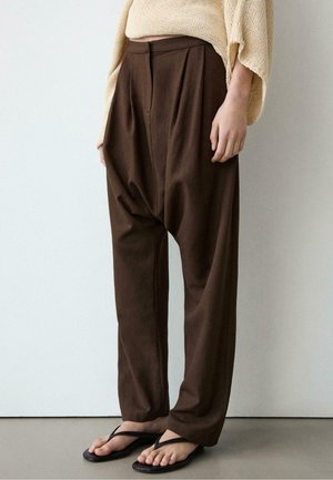 Braune weit geschnittene Hose mit niedrigem Schritt, getragen mit beigem Stricktop und schwarzen Flip-Flops, stehend vor einer einfarbig hellen Wand.