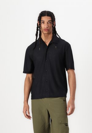 Chemise noire à manches courtes avec un col, présentant un design texturé et des boutons à l'avant, associée à un pantalon cargo olive détendu.