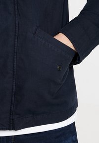 Marinblå jacka med en taktil yta, med en sidoficka med en synlig knapp och en avslappnad passform. Lagd över en vit skjorta.