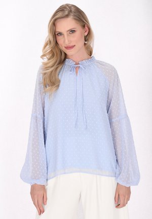 Blusa azzurro chiaro con pois, caratterizzata da un colletto arricciato, dettaglio con fiocco e maniche lunghe trasparenti. Tessuto morbido e strutturato.
