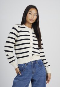 Donna con lunghi capelli scuri indossa un maglione a righe bianche e nere con bottoni e jeans blu, posando con una mano in tasca su uno sfondo semplice.