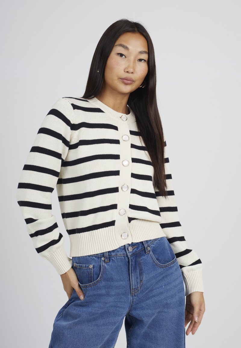 Donna con lunghi capelli scuri indossa un maglione a righe bianche e nere con bottoni e jeans blu, posando con una mano in tasca su uno sfondo semplice.