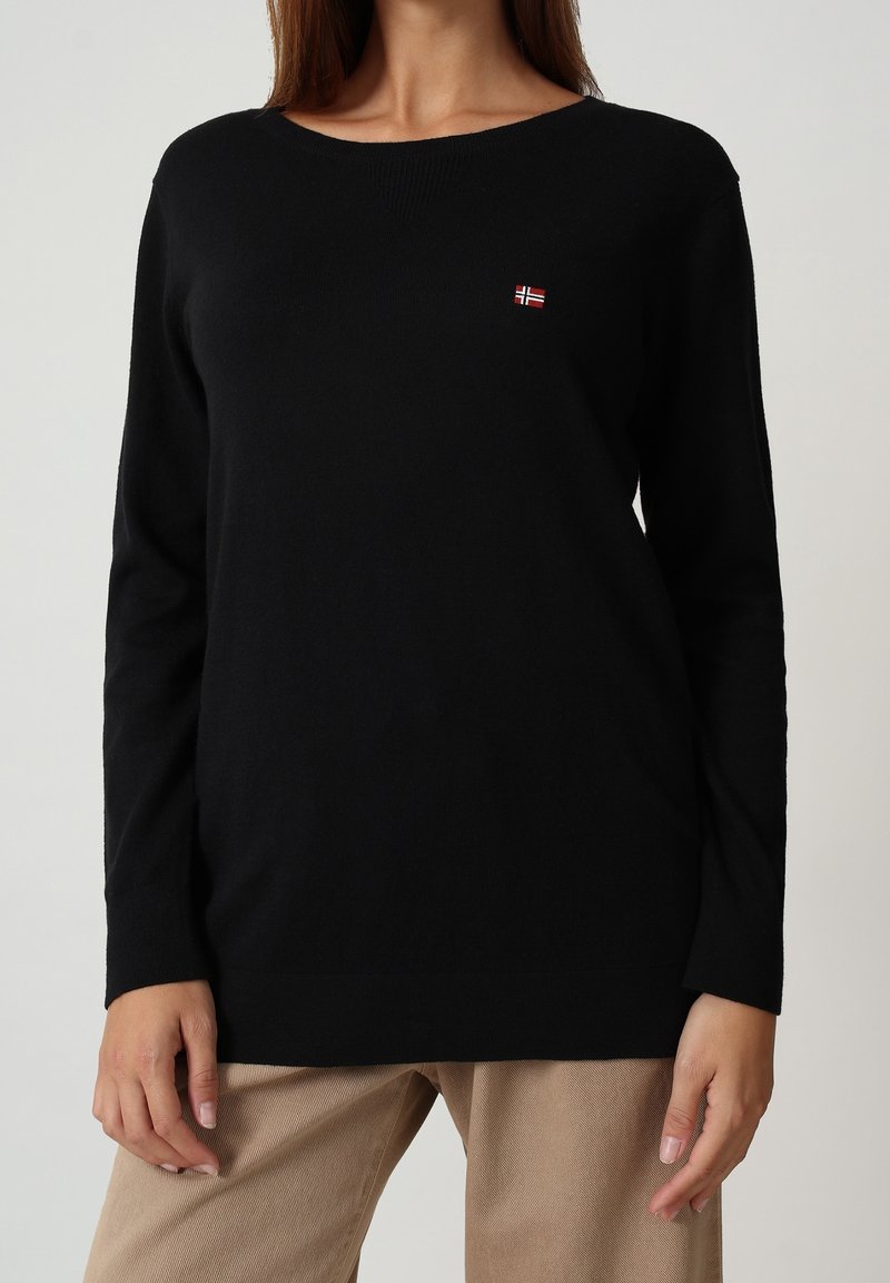 Femme portant un pull noir uni à manches longues avec un petit emblème du drapeau norvégien sur la poitrine et un pantalon beige.