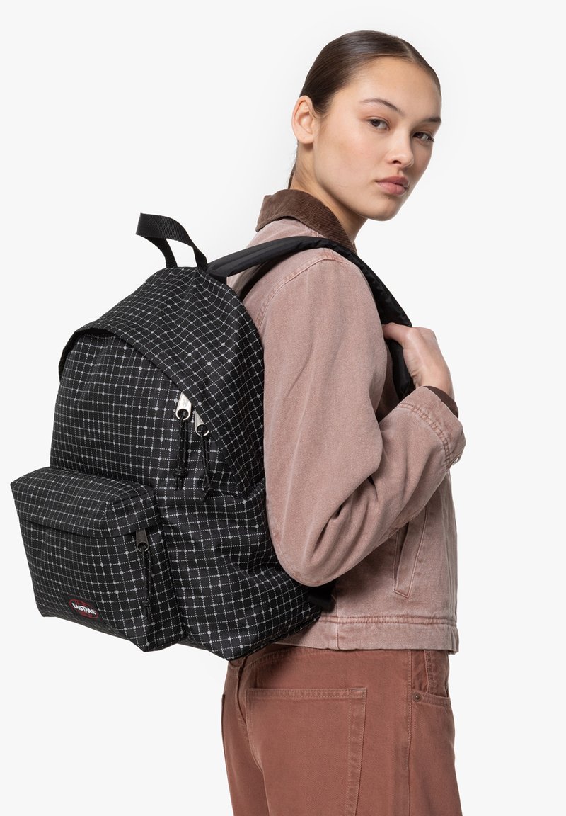 Mochila de cuadros en blanco y negro, material de tela, tapa superior con cierre de cremallera, bolsillo frontal, correas ajustables, detalle de logo sutil.