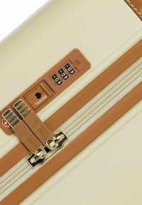 Maleta color crema con una cremallera marrón, que cuenta con un candado TSA, diales de combinación y una superficie texturizada. Los acentos incluyen herrajes de metal.