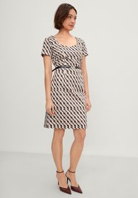Kleid mit kurzen Ärmeln, quadratischem Ausschnitt und einem schwarzen geometrischen Muster auf hellem Hintergrund. Die Taille wird von einem schwarzen Gürtel betont.