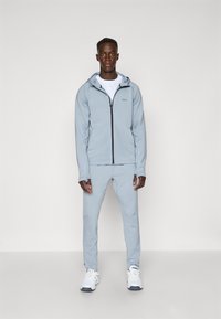 Ensemble de vêtements de sport gris clair composé d'un sweat à capuche zippé et d'un pantalon assorti. Le tissu est lisse, avec une coupe décontractée et des baskets blanches.