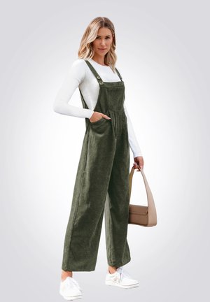 Olijfgroene corduroy jumpsuit met wijde pijpen, verstelbare banden en voorzakken, gecombineerd met een wit longsleeve shirt en sneakers.