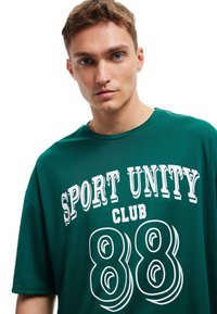 Joven con cabello corto castaño que lleva una camiseta verde del "Sport Unity Club 88", mirando hacia adelante sobre un fondo blanco.