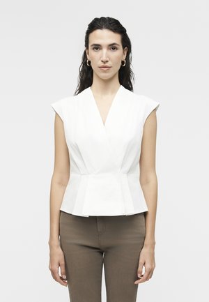 Vrouw met lang, donker haar draagt een mouwloos wit peplumtopje en bruine broek, kijkt recht vooruit tegen een effen grijze achtergrond.