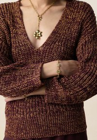 Persoon draagt een paarse en gouden gebreide trui met een gouden bloemhanger aan een ketting en bijpassende gouden armband, armen gekruist.