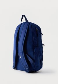 Σακίδιο πλάτης σε ναυτικό μπλε με επενδεδυμένους ιμάντες ώμου, λαβή στο πάνω μέρος, πλαϊνή τσέπη και εμφανείς φερμουάρ πάνω σε απλό ανοιχτό φόντο.