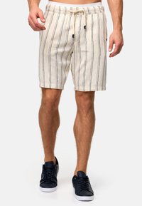 Shorts rayés beige avec des lignes verticales noires, taille avec cordon de serrage et coupe décontractée. Associés à des baskets navy avec des semelles blanches.