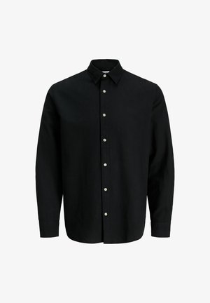 Garçon aux cheveux en bataille portant une chemise noire boutonnée, un pantalon beige et des baskets blanches, debout devant un fond clair uni.