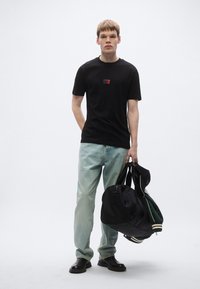 Μαύρο βαμβακερό μπλουζάκι με μικρό κόκκινο λογότυπο, ανοιχτό μπλε τζιν και μαύρα παπούτσια. Κρατάει μαύρο σακίδιο και πράσινο σακάκι.