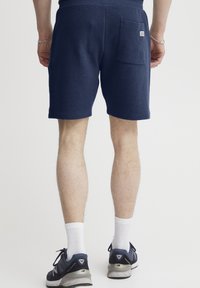 Sötétkék, textured anyagból készült rövidnadrág, hátoldali zsebbel és egy kis márkacímkével. Sportcipővel és fehér zoknival viselve.