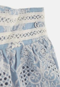 Lichtblauwe stoffen top met ingewikkelde witte borduursels, voorzien van kantafwerking, decoratieve accenten en een zilveren "G" detail.