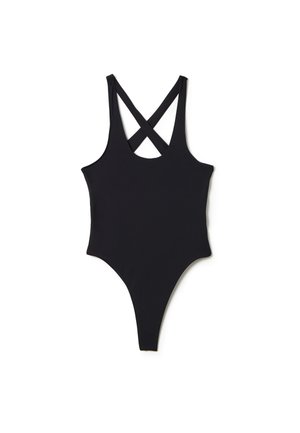 Femme aux longs cheveux foncés, debout face à l'avant, portant un maillot de bain noir taille haute et des bottes noires à lacets devant un mur en béton gris.