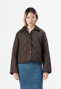 Joven con el pelo a la altura de los hombros que lleva una chaqueta acolchada marrón oscura y una falda de denim azul, de pie contra un fondo blanco.