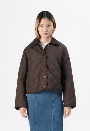 Une jeune personne avec des cheveux à mi-longueur porte une veste matelassée marron foncé et une jupe en denim bleue, se tenant devant un fond blanc.