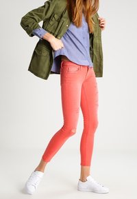 Mujer con pantalones ajustados de color coral, zapatillas blancas, camisa de botones azul y chaqueta verde, ajustándose la chaqueta mientras está de pie contra un fondo sencillo.