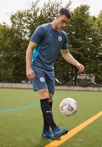 Sporta apģērbs ar tumši zilu krekls ar tirkīza akcentiem un atbilstošām šortiem, melni ceļgala garuma zeķes un zilas sporta kurpes, kopā ar futbola bumbu.