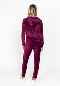 Burgundin väriin kuuluva veluurinen treenipuku, jossa on huppari ja kapenevat housut. Silkkinen pinta, joustavat hihansuut ja yksityiskohtaiset ompeluakoristukset.