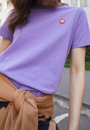 Femme portant un t-shirt lilas avec un petit logo rond sur la poitrine et un pull marron noué autour de la taille en extérieur.